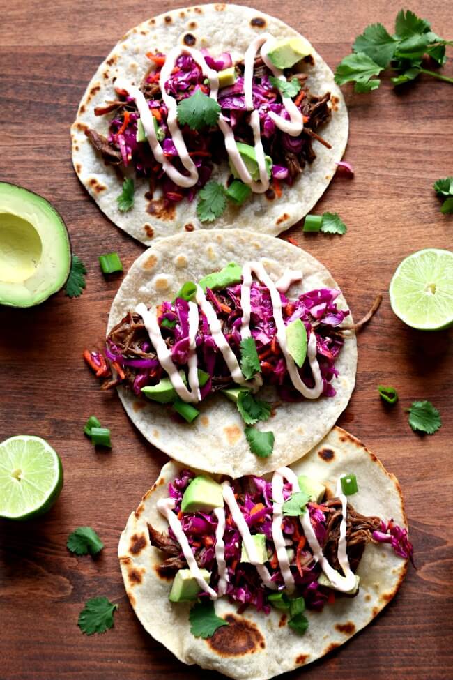 Pressure Cooker/Slow Cooker Korean Beef Tacos--tender shredded seasoned beef is rolled up in a warm tortilla with crunchy cabbage, diced avocado, chopped cilantro, sriracha sour cream and a splash of roasted garlic seasoned rice vinegar. When you bite into this taco it's like a party in your mouth. All the flavors and textures come together in a perfect union.